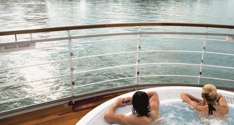Couple se détendant dans un jacuzzi extérieur sur le pont d'un bateau de croisière fluviale avec l'eau scintillant à côté d'eux.