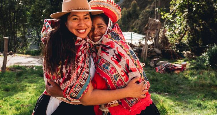 Dos mujeres sonrientes con vestimenta andina vibrante se abrazan en un entorno rural.