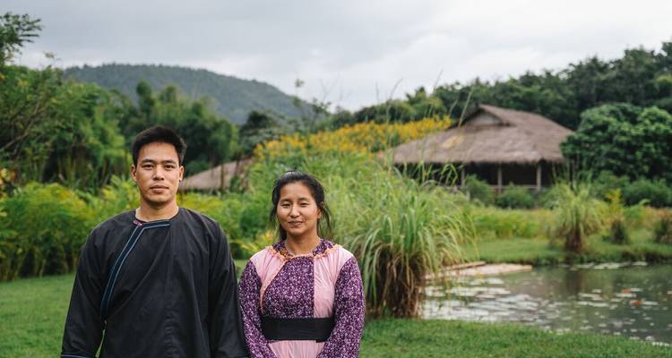 Un homme et une femme vêtus de costumes traditionnels se tiennent devant des écolodges au toit de chaume et des étangs.
