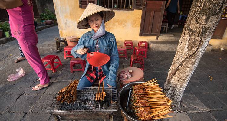 Jeune vendeur portant un chapeau conique qui ravive les brochettes grillant sur un barbecue de rue au charbon de bois.