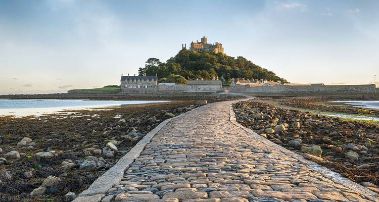Calzada de adoquines que emerge con la marea baja, llevando al castillo medieval en la cima del Mont Saint Michel bajo una luz dorada.