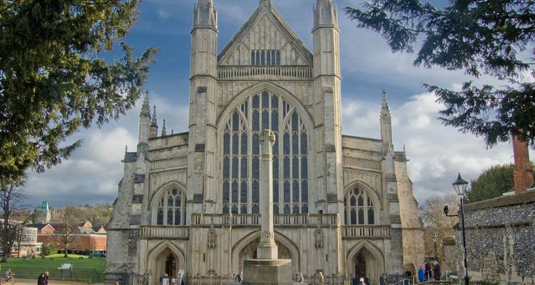 La gran fachada occidental de la Catedral de Winchester enmarcada por árboles bajo un cielo parcialmente nublado.