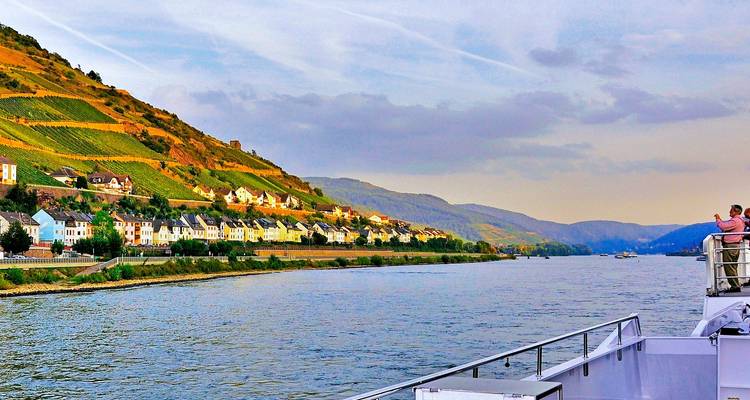 Vine-covered hills and pastel villages line the Rhine as passengers enjoy the view from a cruise ship deck.
