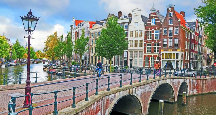 Cyclists cross a classic brick bridge over an Amsterdam canal lined with gabled houses and trees.