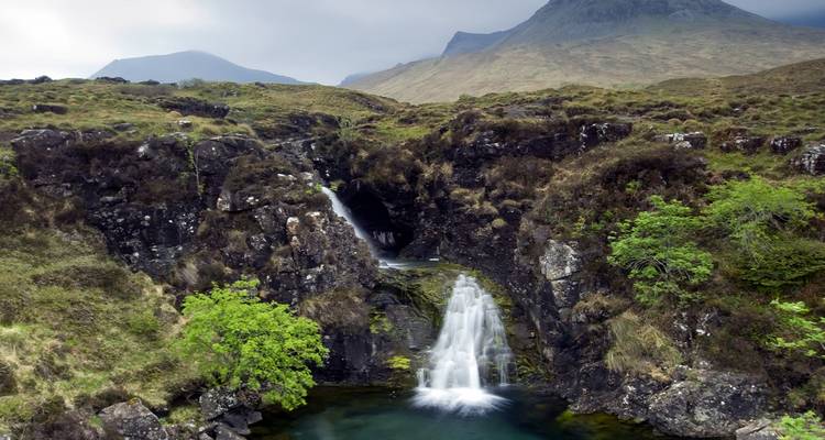 Cascade d'eau tombant dans un petit bassin avec des montagnes en arrière-plan.