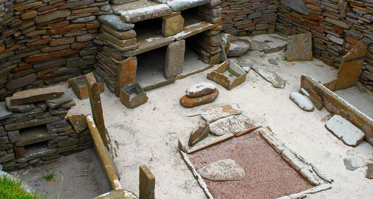 Overblijfselen van een stenen huis uit het Neolithicum van Skara Brae met haarden en stenen meubels op een zandige vloer.