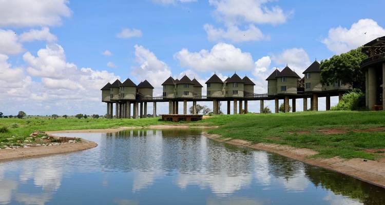 Row of stilted lodge huts reflected in a calm water channel under a blue sky with puffy clouds.