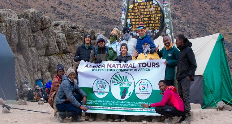 Groot trekkingteam dat een spandoek toont bij een berghutbord met tenten en rotsachtige achtergrond