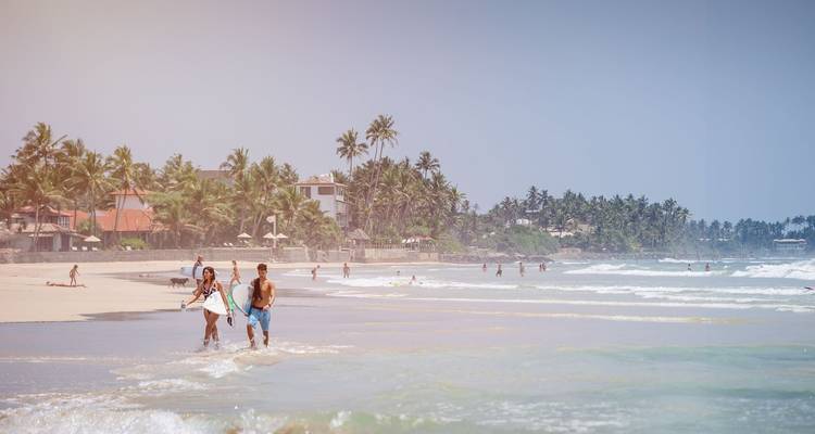 Menschen, die entlang eines sandigen Strandes mit Palmen spazieren gehen.