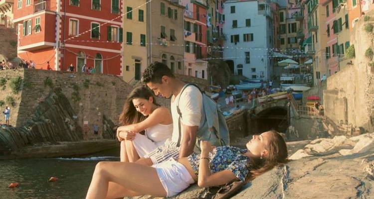 Three friends relaxing on sun-warmed rocks beside colorful cliffside houses of Cinque Terre.