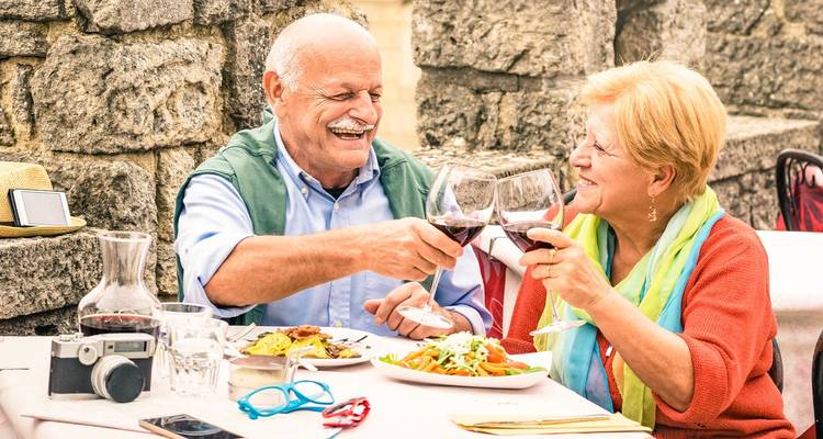 Lachendes älteres Paar stößt mit Gläsern Rotwein über Tellern mit Pasta in einem Außenbereich eines Restaurants mit Steinmauern an.