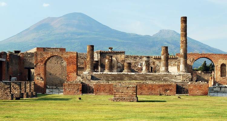 Alte Ziegelstein- und Steinruinen von Pompeji mit der konischen Silhouette des Berges Vesuv, der sich im Hintergrund unter einem klaren Himmel erhebt.