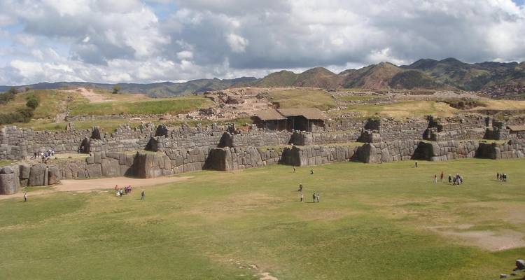 Weitläufiger Grasbewuchs umgebener Platz, der von massiven Steinmauern von Saqsaywaman umgeben ist, mit Besuchern, die umhergehen.