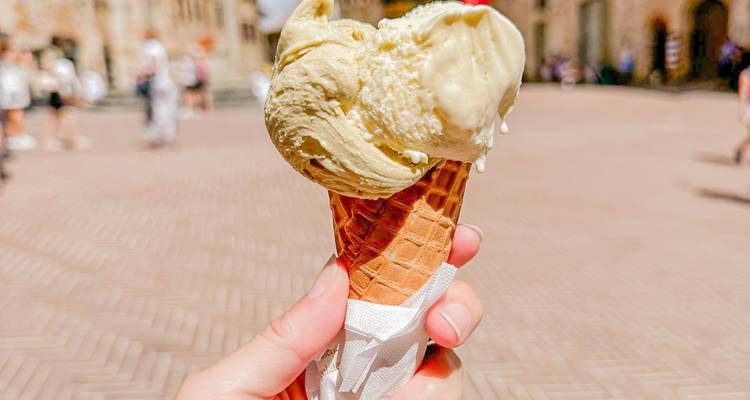 Primer plano de una mano sosteniendo un cono de gelato cremoso con una plaza medieval borrosa de fondo.