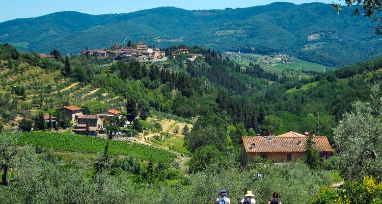 Vue panoramique de la campagne toscane ondulante avec des randonneurs descendant vers un village perché sur une colline.