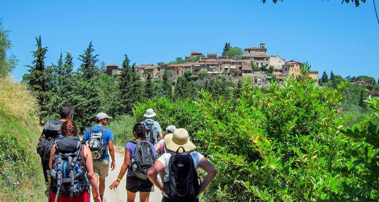 Des randonneurs chargés de sacs à dos suivent un sentier de terre en direction d'un village toscan perché sur une colline sous un ciel bleu dégagé.