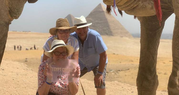 Tourists crouching under camel legs with pyramids in the background.