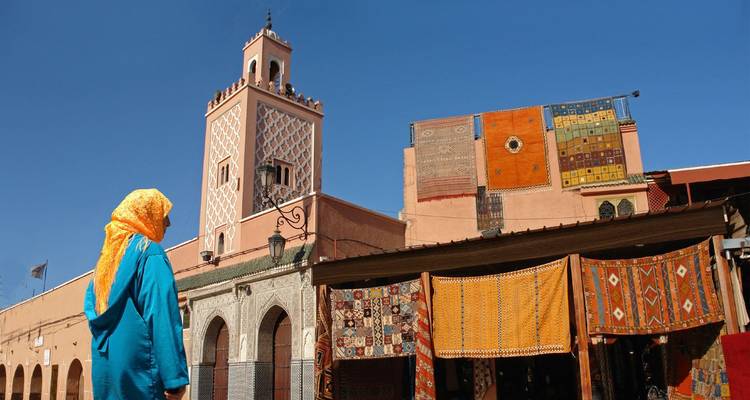Lokale vrouw in blauwe robe kijkt uit naar een minaret boven kleurrijke tapijtkramen in de souk van Marrakesh.