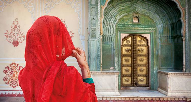 A woman in a red sari standing before an ornate Indian doorway.