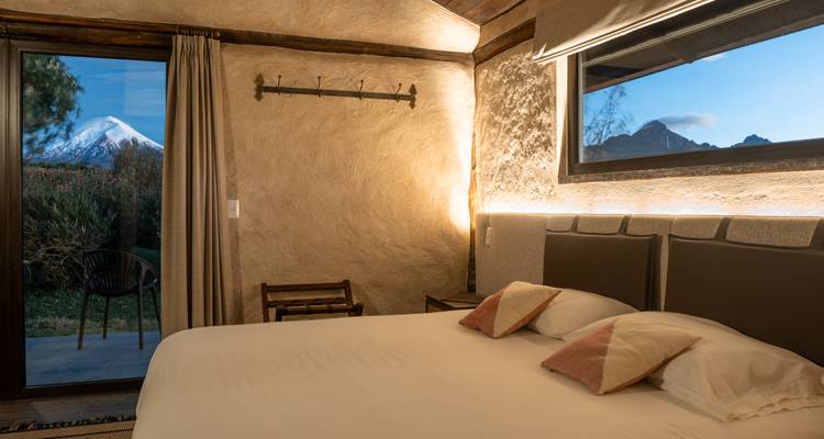 Gezellig hotelkamertje met warm licht en een groot raam met uitzicht op een sneeuwbedekte vulkaan.