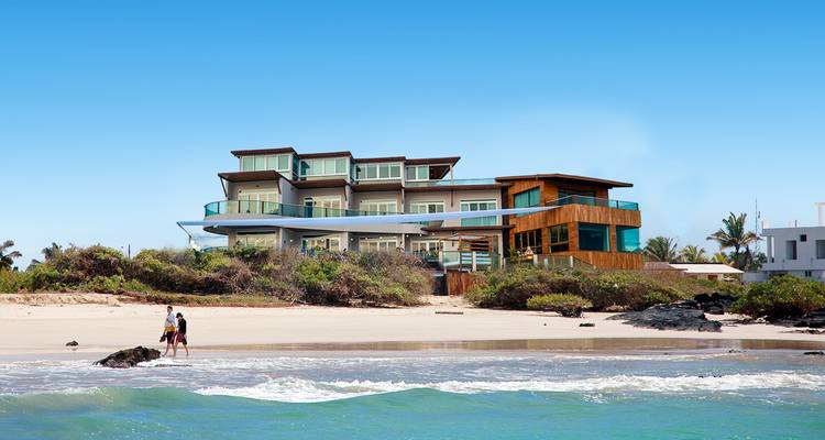 Complexe hôtelier moderne en front de mer avec balcons donnant sur la mer turquoise et un couple marchant sur le rivage sablonneux.