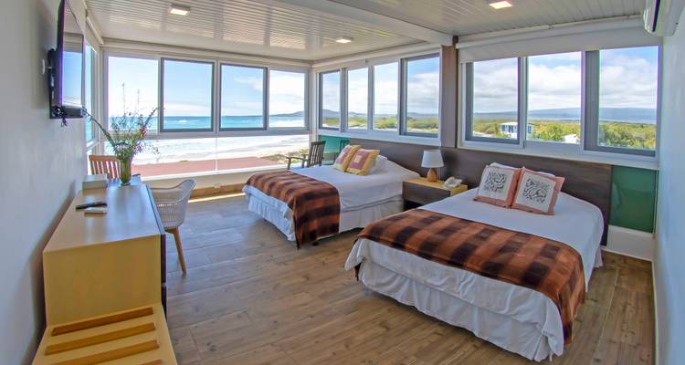 Chambre d'hôtel lumineuse avec de grandes fenêtres donnant sur l'océan et la plage de sable, deux lits jumeaux et décoration moderne.