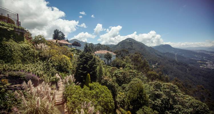 Sendero en un exuberante jardín y villas en la ladera con vistas a montañas boscosas y al cielo.