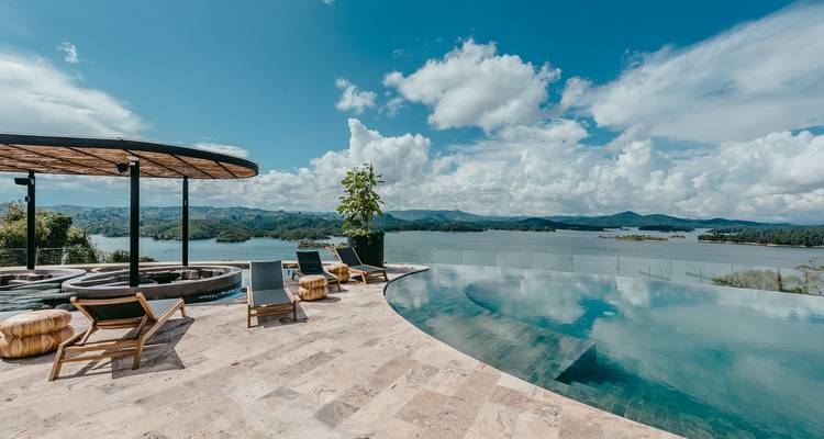 Piscina infinita y tumbonas con vistas a un lago reluciente y a colinas verdes lejanas bajo cielos despejados.