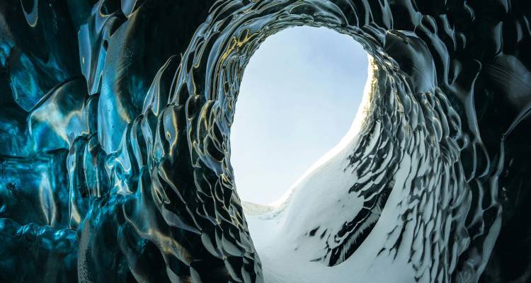 Dentro de una cueva de hielo, paredes azul-negras de glaciar esculpido enmarcan una abertura ovalada hacia el cielo brillante.