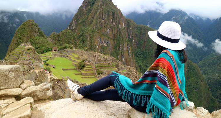 Eine Person, die auf einem Felsen sitzt und auf Machu Picchu blickt, trägt einen bunten Poncho.