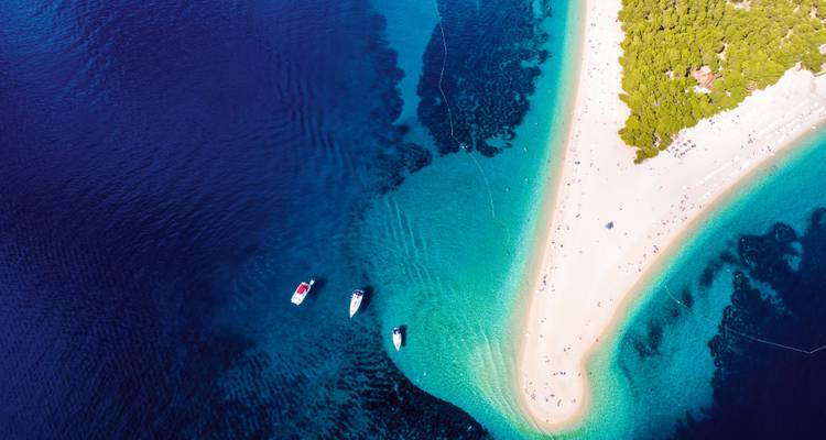 Toma aérea con dron de gran angular de la lengua de arena de Zlatni Rat extendiéndose hacia el agua de color zafiro con barcos fondeados y densos pinos cerca.