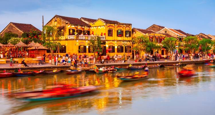 Historische gelbe Gebäude säumen den Fluss in Hoi An, während unscharfe Longtail-Boote bei Sonnenuntergang über das Wasser gleiten.