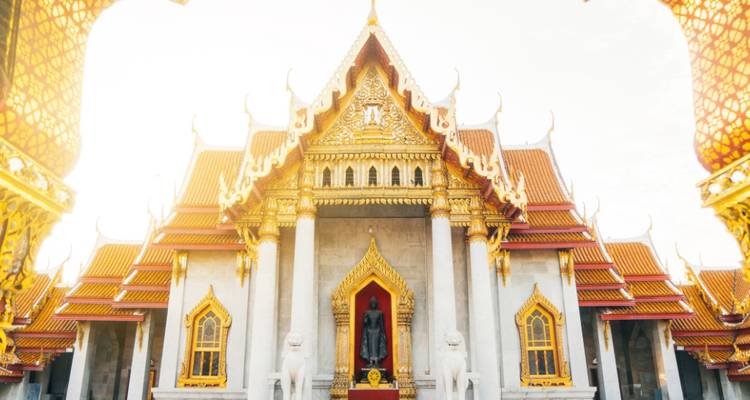 Templo de Mármol brillantemente iluminado en Bangkok con frontones ornamentales y detalles dorados contra un cielo pálido.