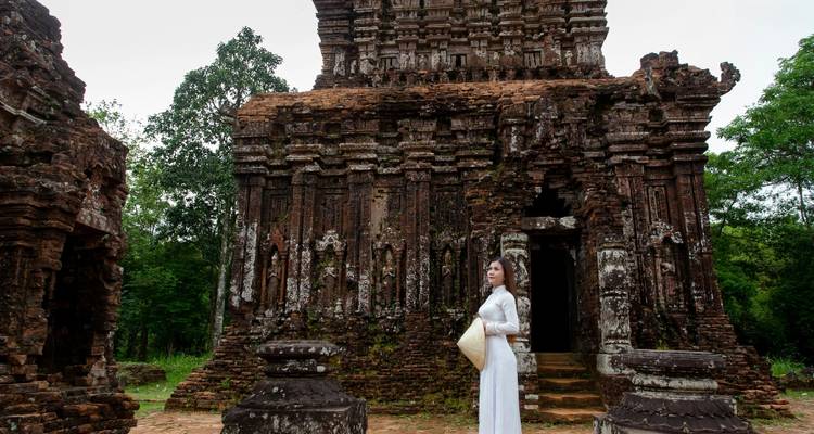 Woman in white ao dai holding a conical hat in front of an ancient Cham tower amid greenery.