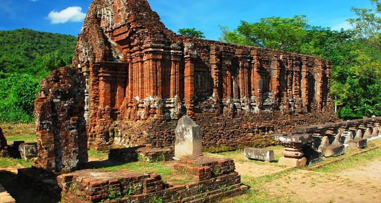Red-brick ruins of an ancient temple complex against a verdant hillside and blue sky.