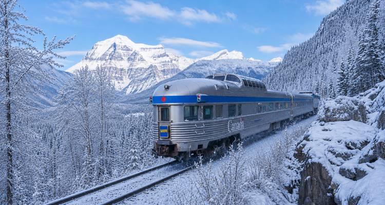 Silver passenger train winding through snowy mountains under a bright blue winter sky