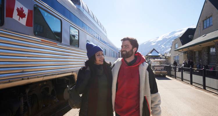 Smiling couple walking along a platform beside a blue Canadian train with snowy mountains behind