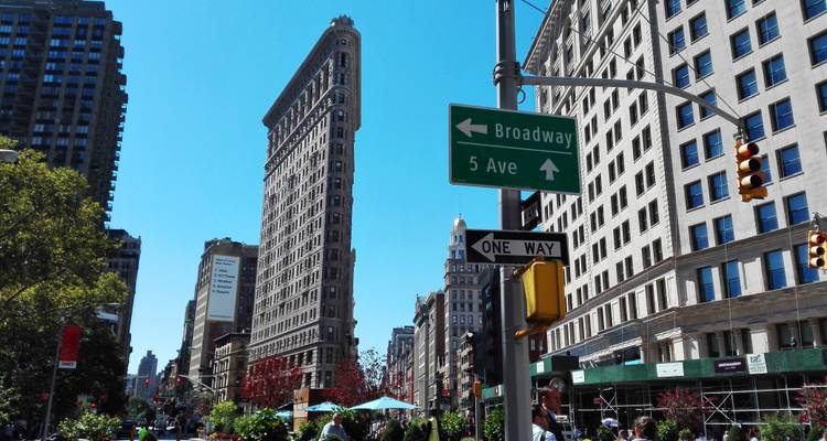 Street-level view of New York’s Flatiron Building with Broadway and 5th Avenue signs, traffic lights and pedestrians on a sunny day.