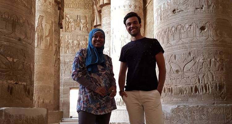Deux jeunes voyageurs se tiennent souriants dans une salle de temple égyptien antique bordée de colonnes ornées couvertes de hiéroglyphes.