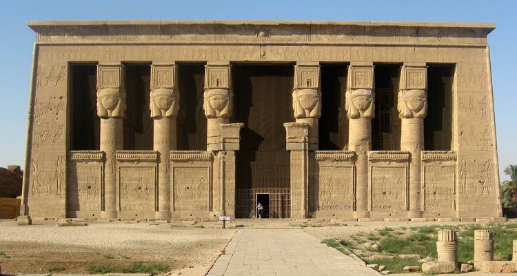 Vue de face de la façade monumentale en grès d'un ancien temple égyptien avec des colonnes à tête d'Hathor sous un ciel bleu dégagé.