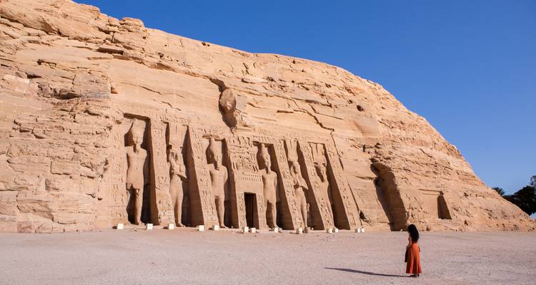 Eine Person, die vor den Abu-Simbel-Tempeln steht, die in eine Felswand gemeißelt sind, bei klarem blauem Himmel.