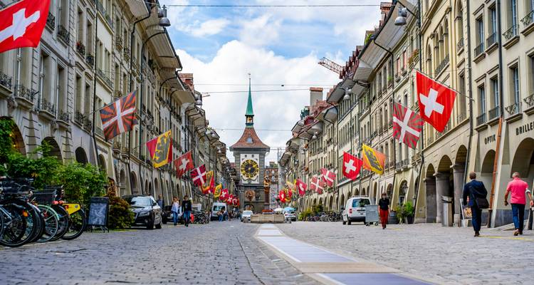 Flag-lined medieval street in Bern leads to the Zytglogge clock tower under bright skies.
