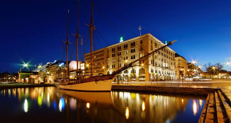 Puerto nocturno en Helsinki con un velero histórico reflejado en aguas tranquilas y edificios iluminados.