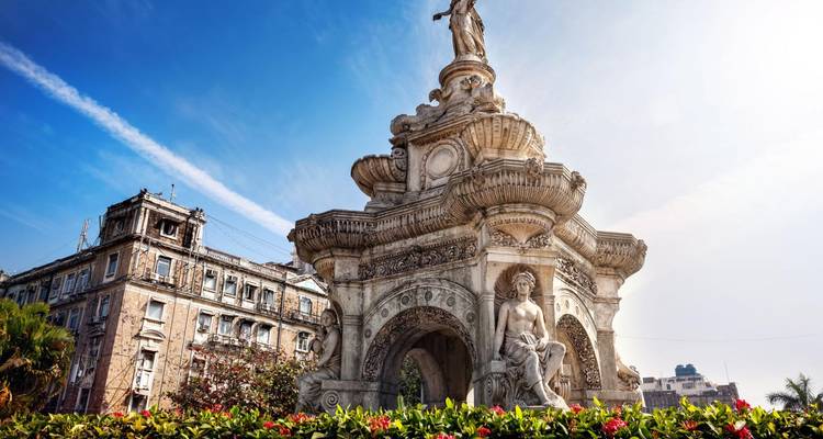 Ornate stone Flora Fountain stands amidst flowers and historic colonial buildings in bright daylight.