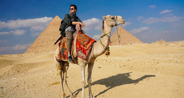 A man sitting on a camel with the pyramids in the background.