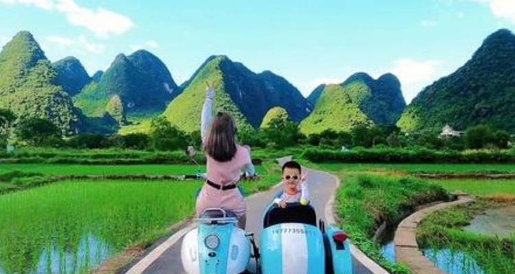 Two people on a scooter in a rural area with hills in the background.