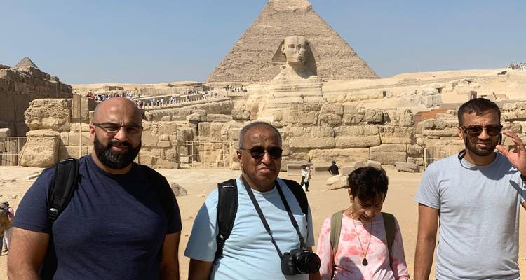 Four tourists pose in front of the Great Sphinx with a pyramid in the background under a clear blue sky.