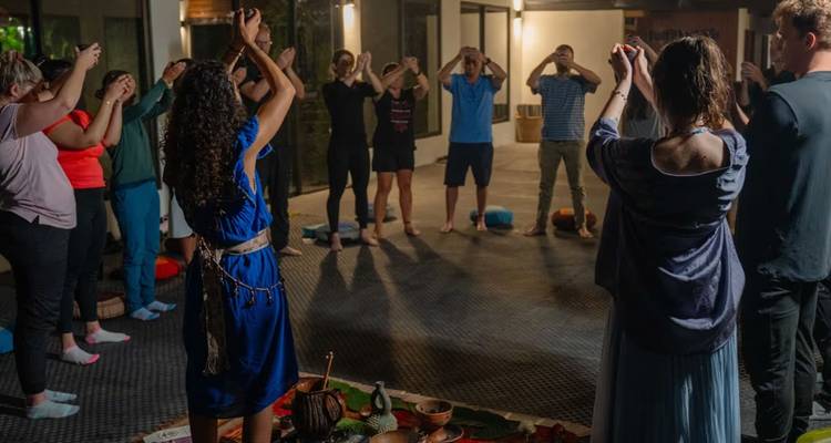Un cercle de participants lève les mains lors d'une cérémonie en soirée à l'intérieur, des tapis et des objets rituels disposés au centre.