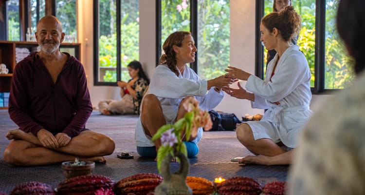Les participants vêtus de robes blanches sont assis en position de lotus à l'intérieur, souriant et se saluant mutuellement lors d'une séance de bien-être.