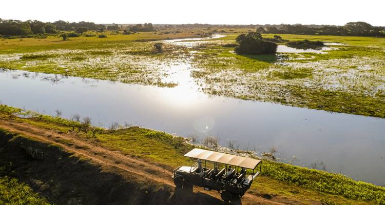 Offenes Safariauto fährt entlang des Randes eines Feuchtgebiets, wobei sich die Sonne im Wasser spiegelt und sich dahinter weite Ebenen erstrecken.